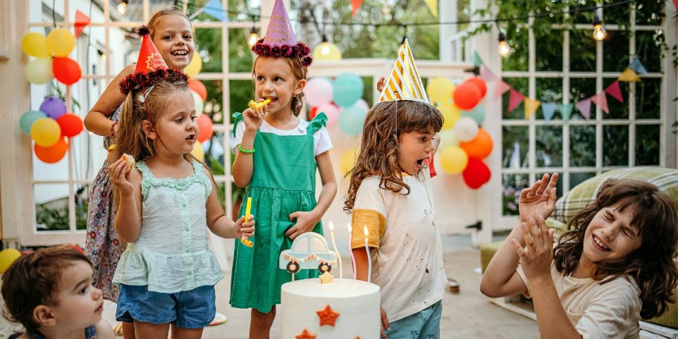 Children with cake standing around table on birthday party in garden in summer.