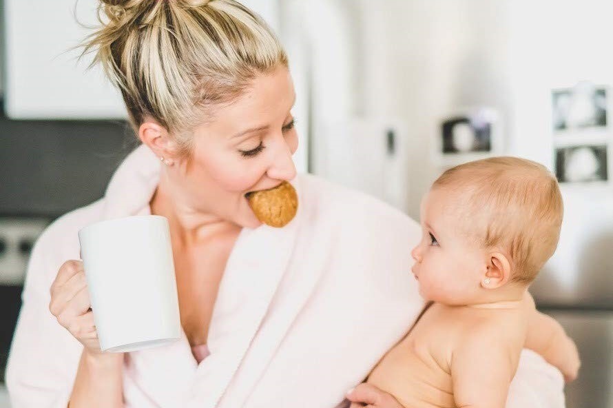 A mom smiling at her baby, coffee in hand, while eating a lactation cookie.